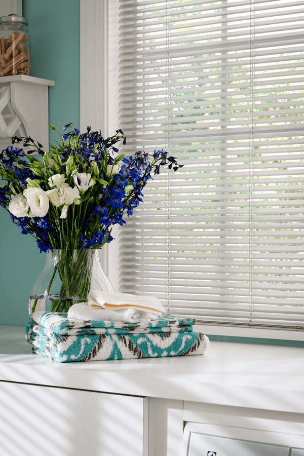 White mini blinds filtering natural light in a bright laundry room with folded towels and a glass vase of fresh flowers.